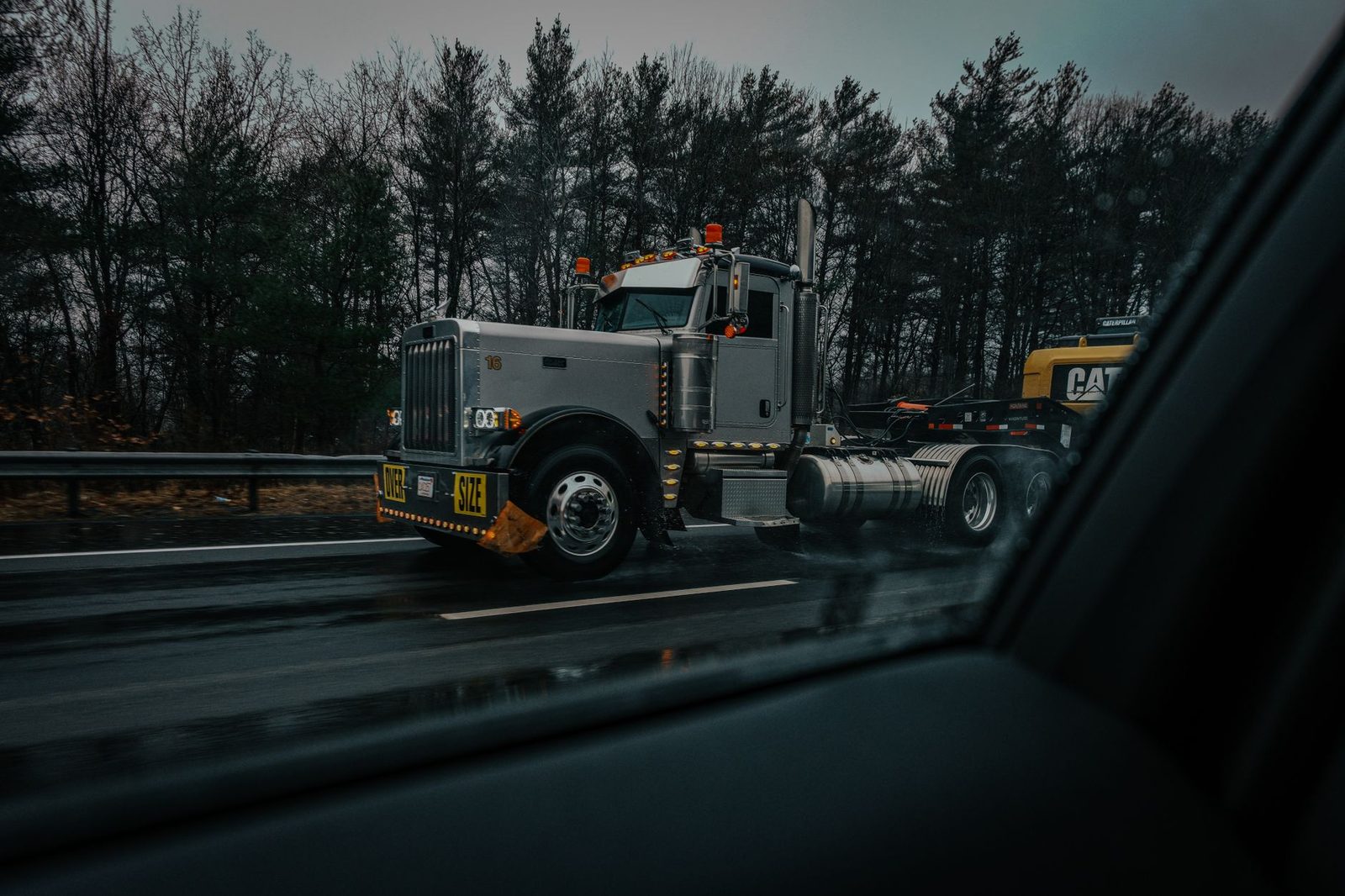 A large truck hauling an oversize load on a rainy highway, seen through a dark, moody car window.