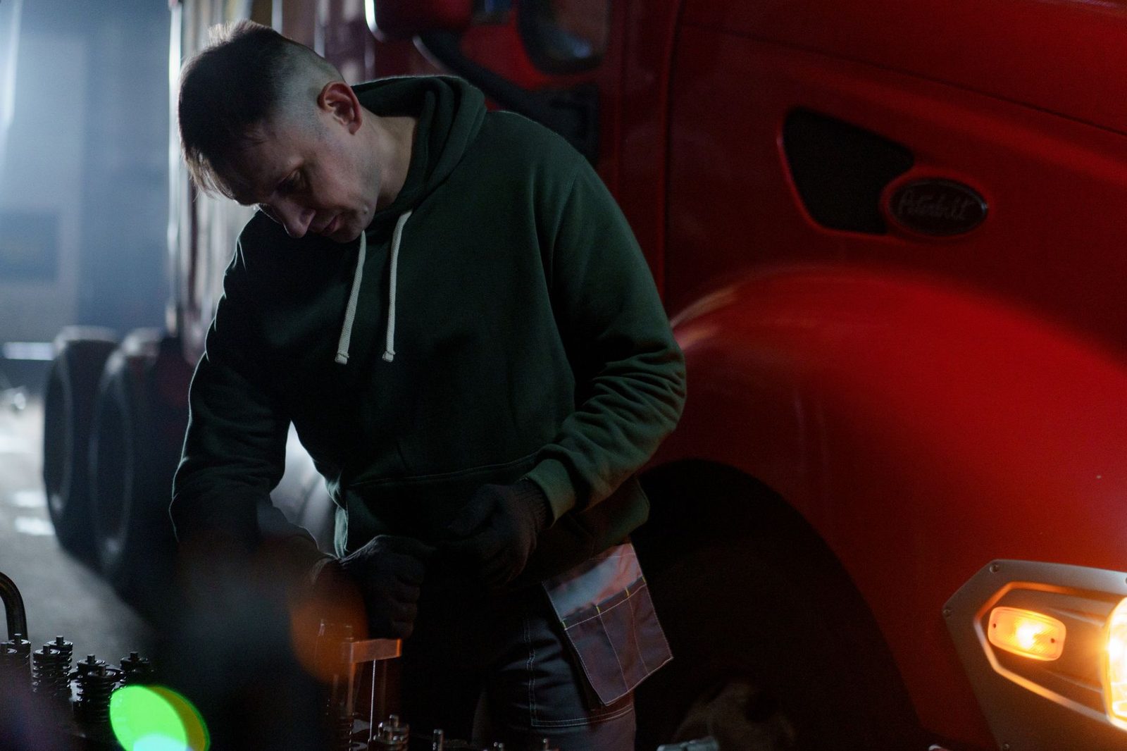 A male mechanic works on a truck engine in a dimly lit garage.