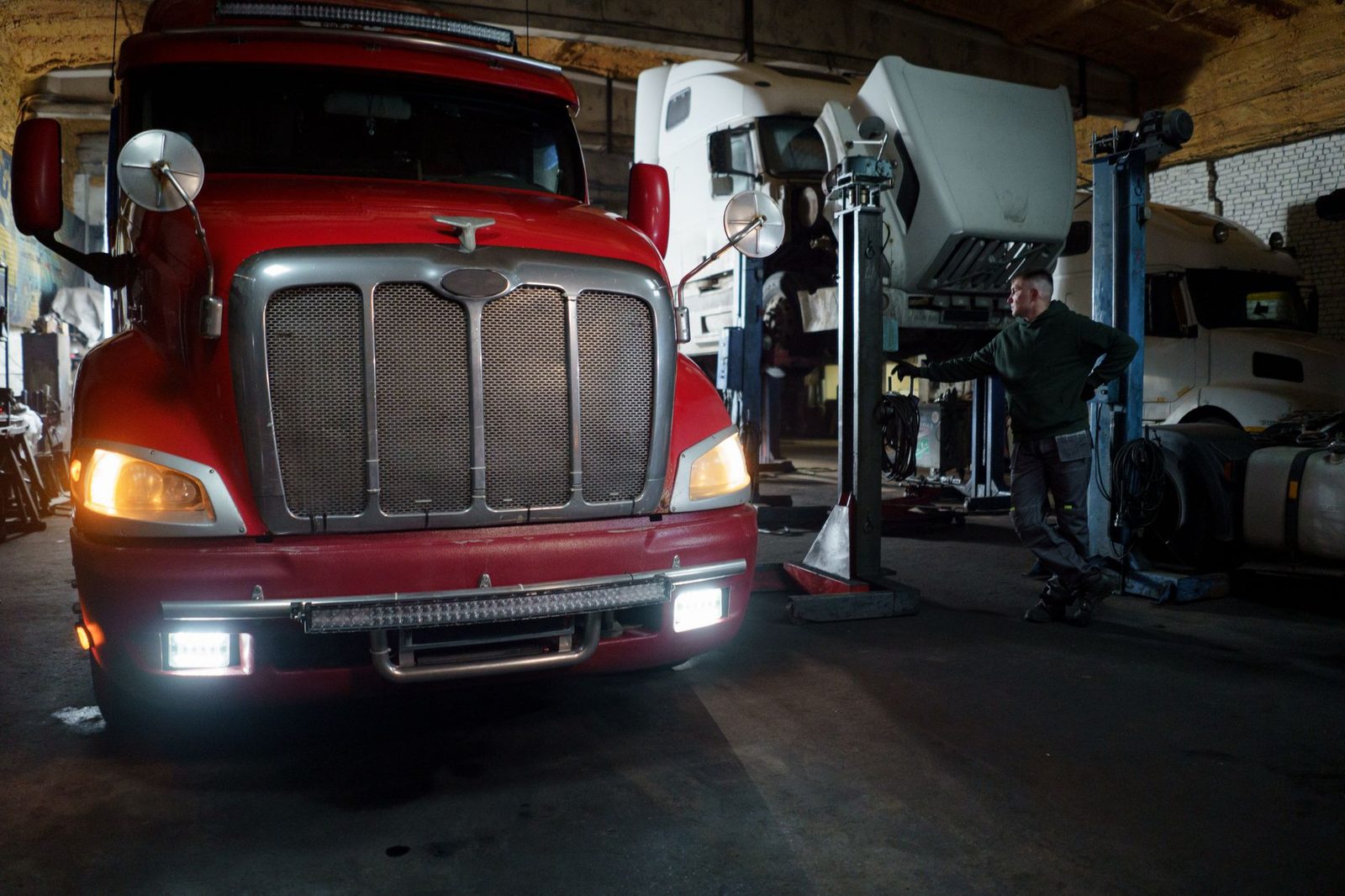 A red truck being serviced by a mechanic in a garage with vehicles raised on lifts.