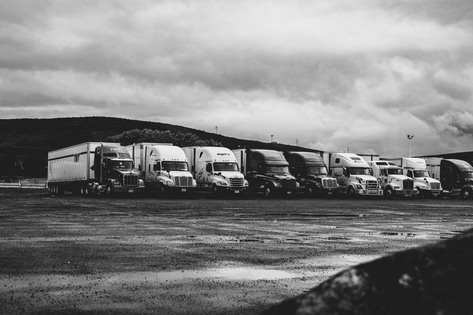 Rows of parked semi trucks sit in a rain-soaked lot in black and white.
